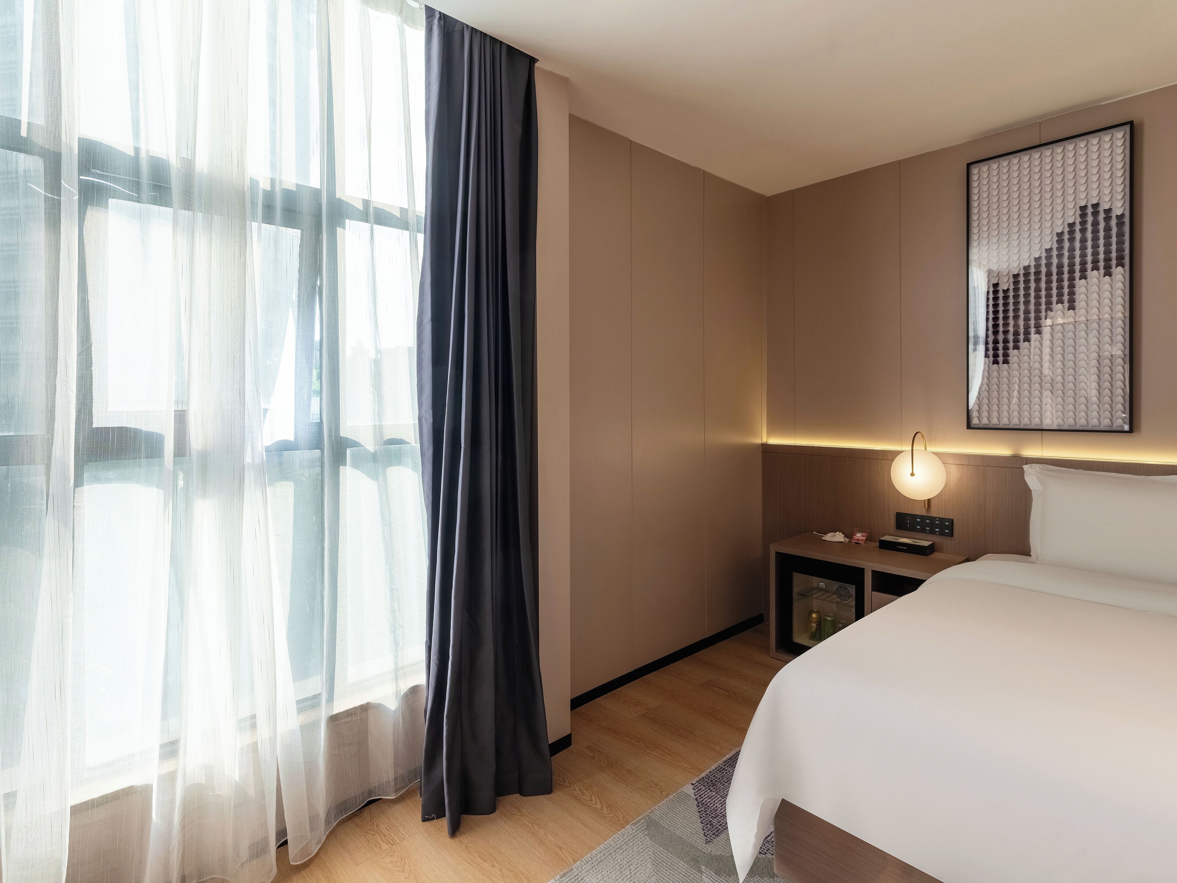 Ein Hotelzimmer mit einem großen Bett, einem Nachttisch mit Lampe, einem gerahmten Kunstwerk an der Wand und transparenten Vorhängen am Fenster.