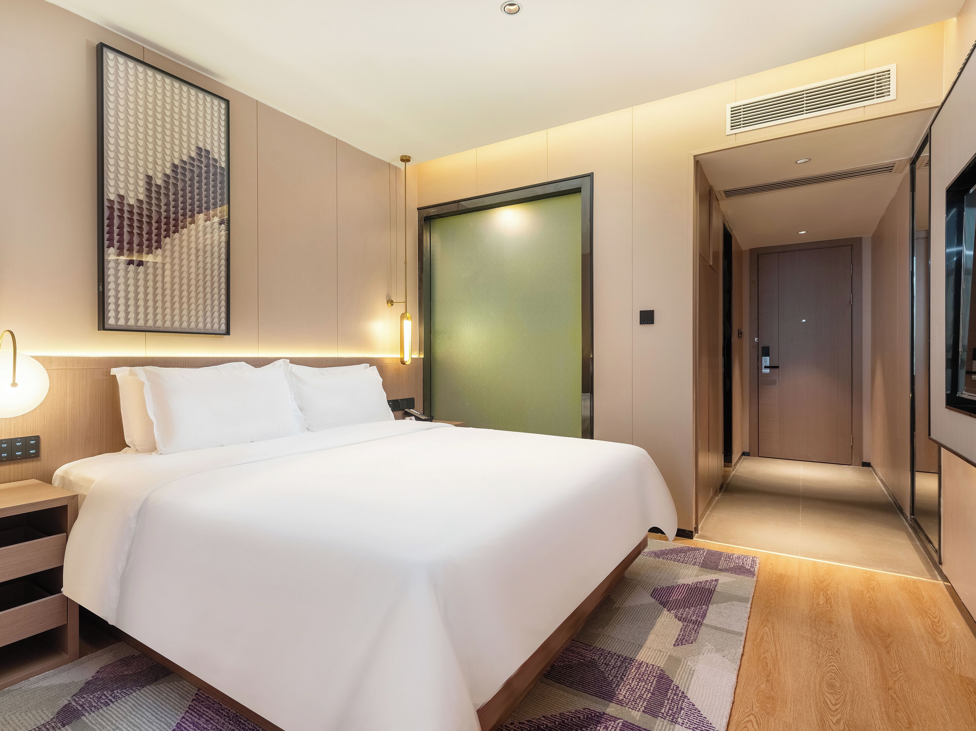 A modern hotel room with a large bed, a bedside table, a wall-mounted artwork, a door, and a hallway.