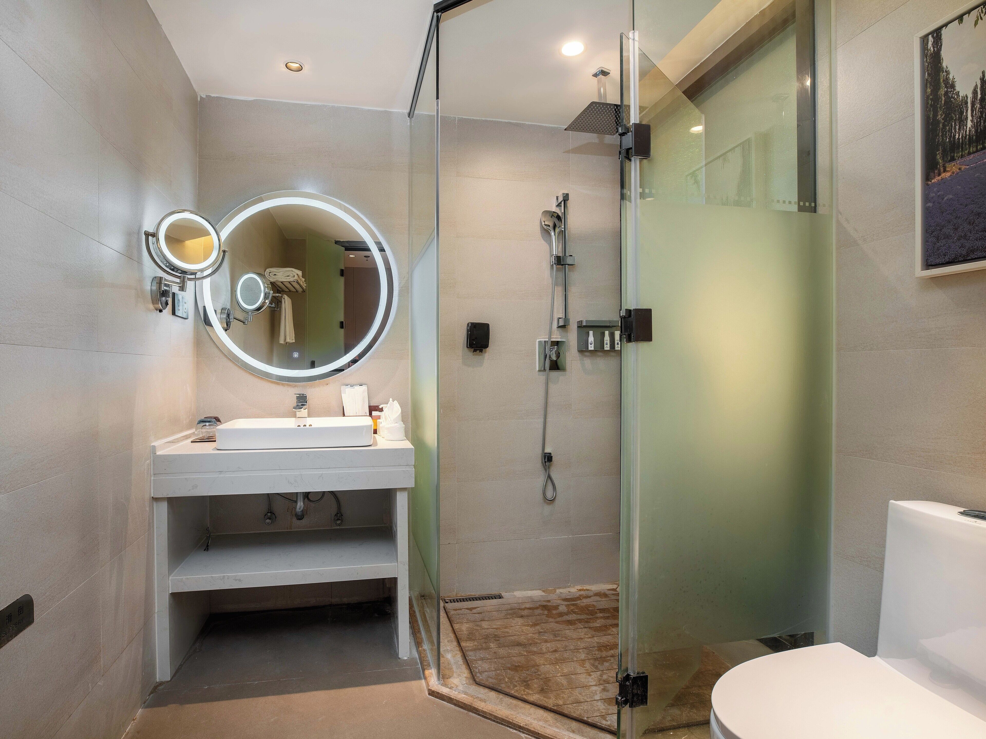Un baño moderno con ducha de vidrio, espejo redondo con iluminación y lavamanos blanco.
