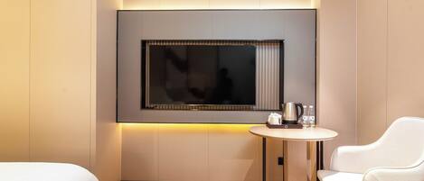 Una habitación de hotel moderna con un televisor de pantalla plana grande montado en la pared, una pequeña mesa redonda con una tetera y botellas de agua, un sofá blanco y una cama bien hecha.