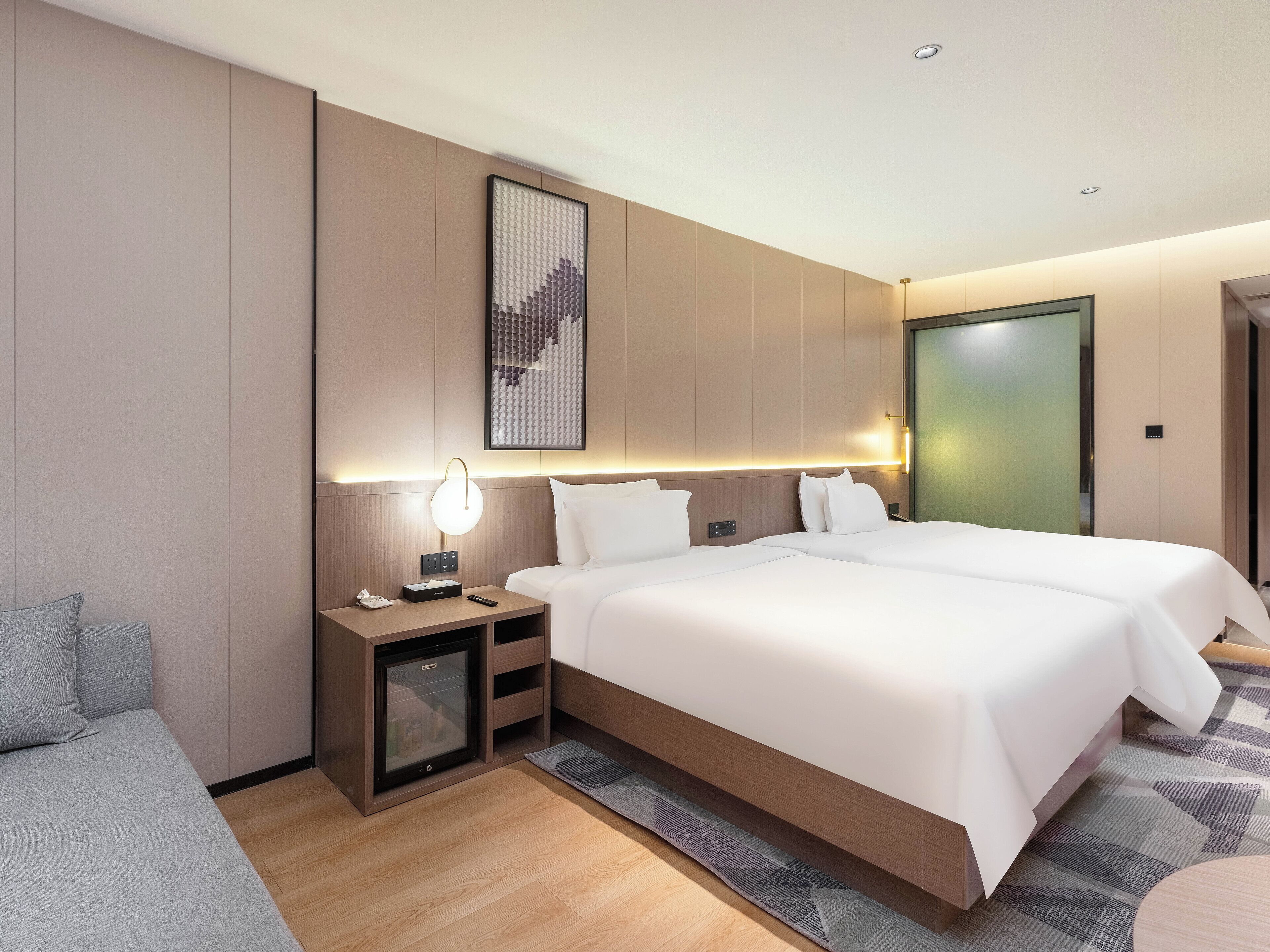 Ein modernes Hotelzimmer mit einem großen Bett, Nachttischen, einem kleinen Kühlschrank und einem an der Wand hängenden Kunstwerk.