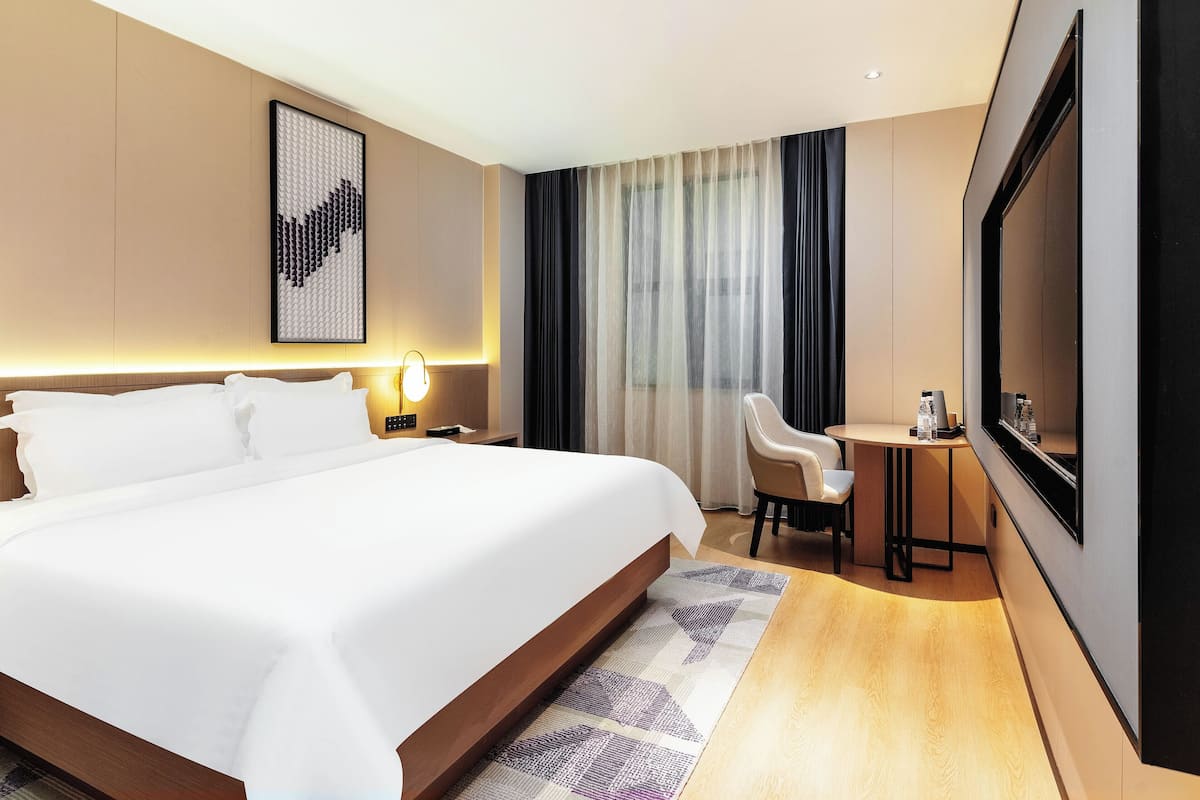 Ein Hotelzimmer mit einem großen Bett, einem Schreibtisch mit Stuhl, einem Fernseher und einem Fenster mit Vorhängen.