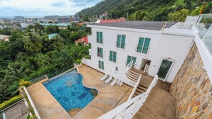 A white building with a swimming pool, surrounded by greenery and a view of a cityscape. - Sea View 2BR Condo Prime Location C236 (Karon)
