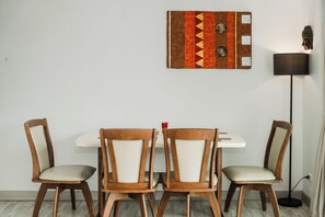 A dining area with a white table and four wooden chairs with cushioned seats. A decorative wall hanging is above the table, and a floor lamp is to the right. - Sea View 2BR Condo Prime Location C236 (Karon)