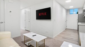 Eine moderne Wohnung mit heller Innenarchitektur, einem Wand-Fernseher, der "NETFLIX" anzeigt, einem kleinen Tisch mit Fernbedienung und einer Küchenzeile.