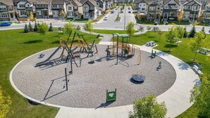 Ein Spielplatz mit Schaukeln und Klettergerüsten, umgeben von einer kreisförmigen Kiesfläche, in einer Wohngegend mit Häusern und blauem Himmel.