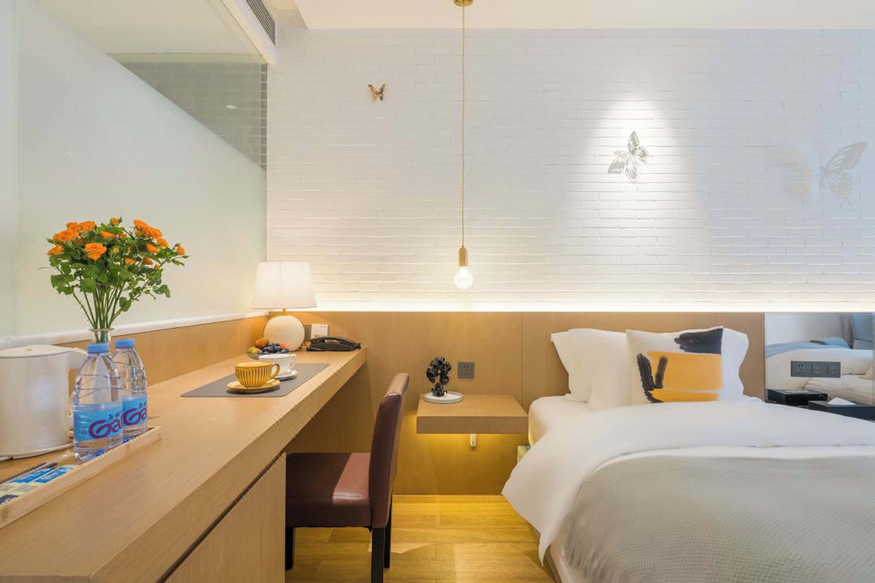 A hotel room with a wooden desk, a chair, a bed with white bedding, a nightstand, a lamp, and a vase with flowers.