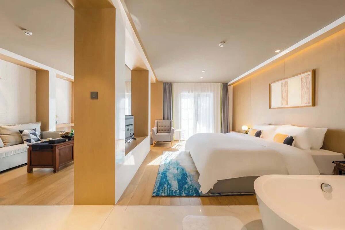 A modern hotel room with a large bed, a sitting area with a sofa and armchair, a desk, and a bathroom with a bathtub.