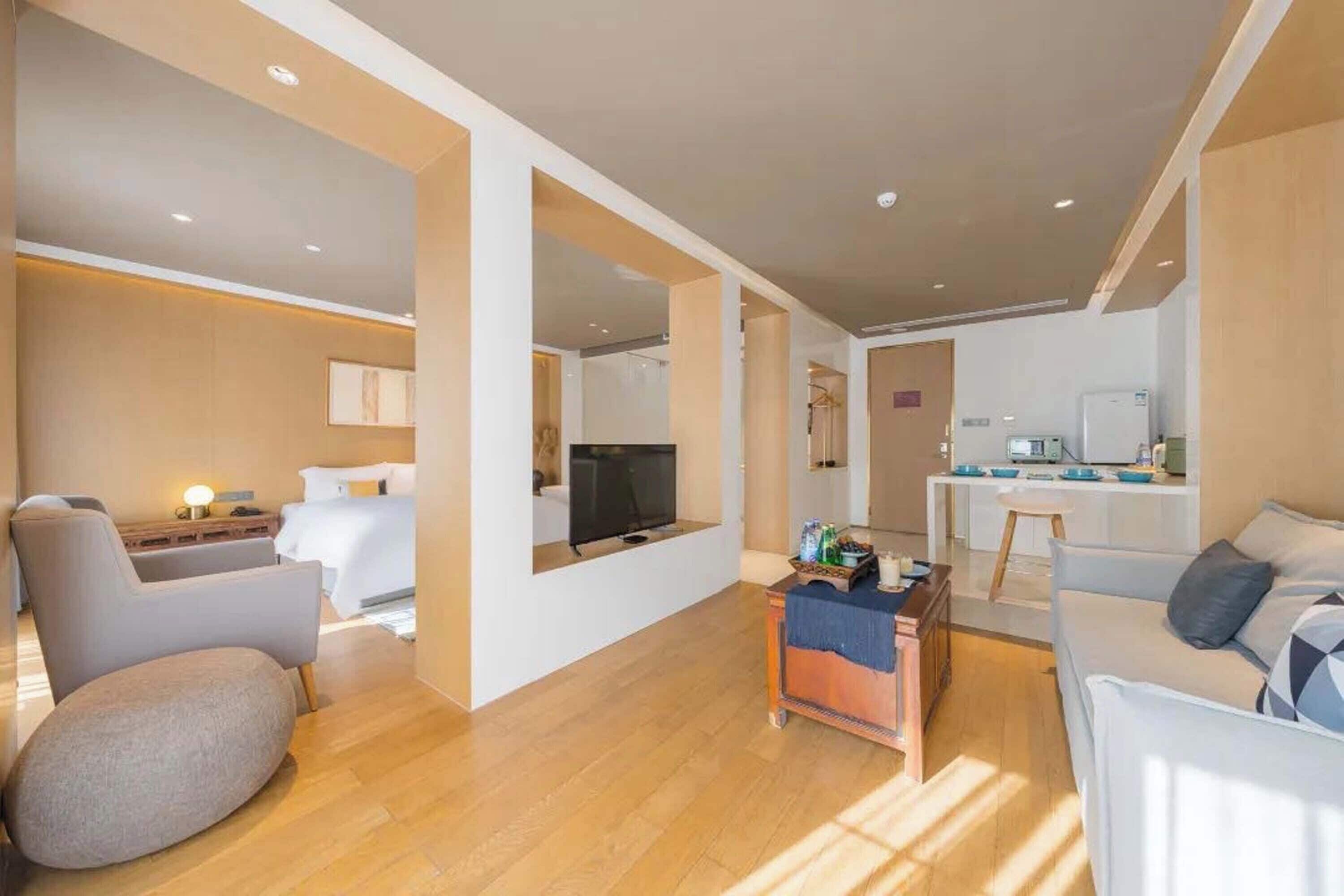 A modern living room with a sofa, armchair, and a small coffee table. There is a TV mounted on the wall and a kitchen area in the background.