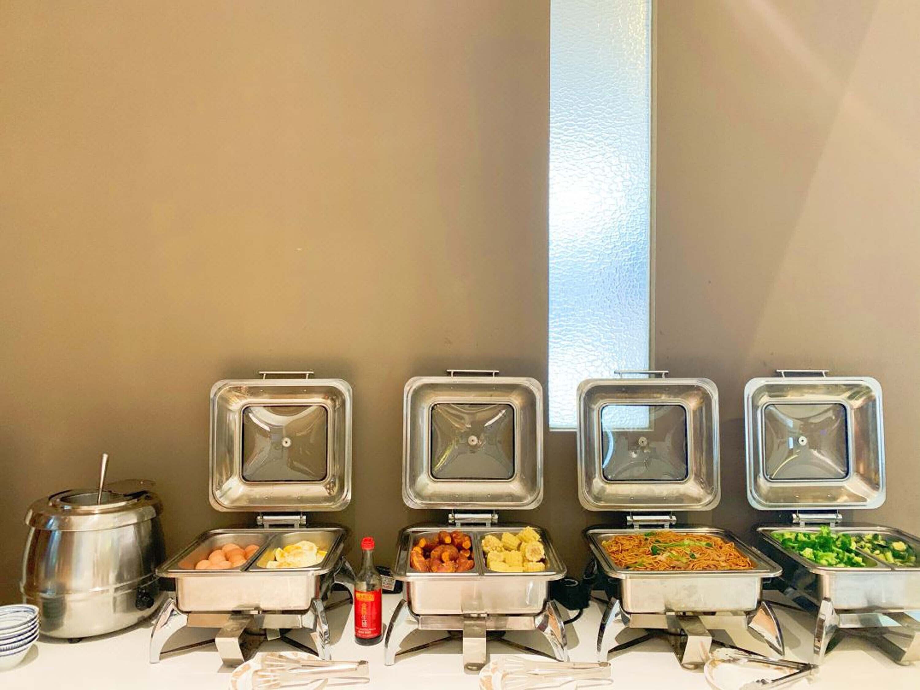 Een buffet met diverse gerechten in metalen bakken, een koffiekan en toevoegingen.