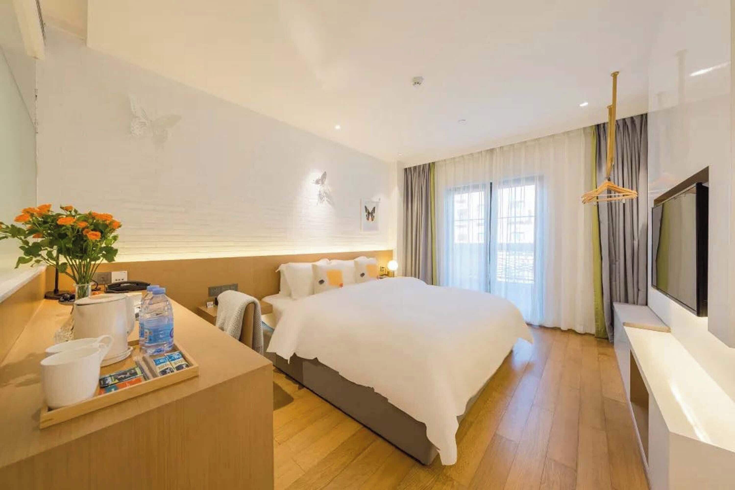 A modern hotel room with a large bed, a bedside table with a tray of breakfast items, a TV, and a large window with curtains.