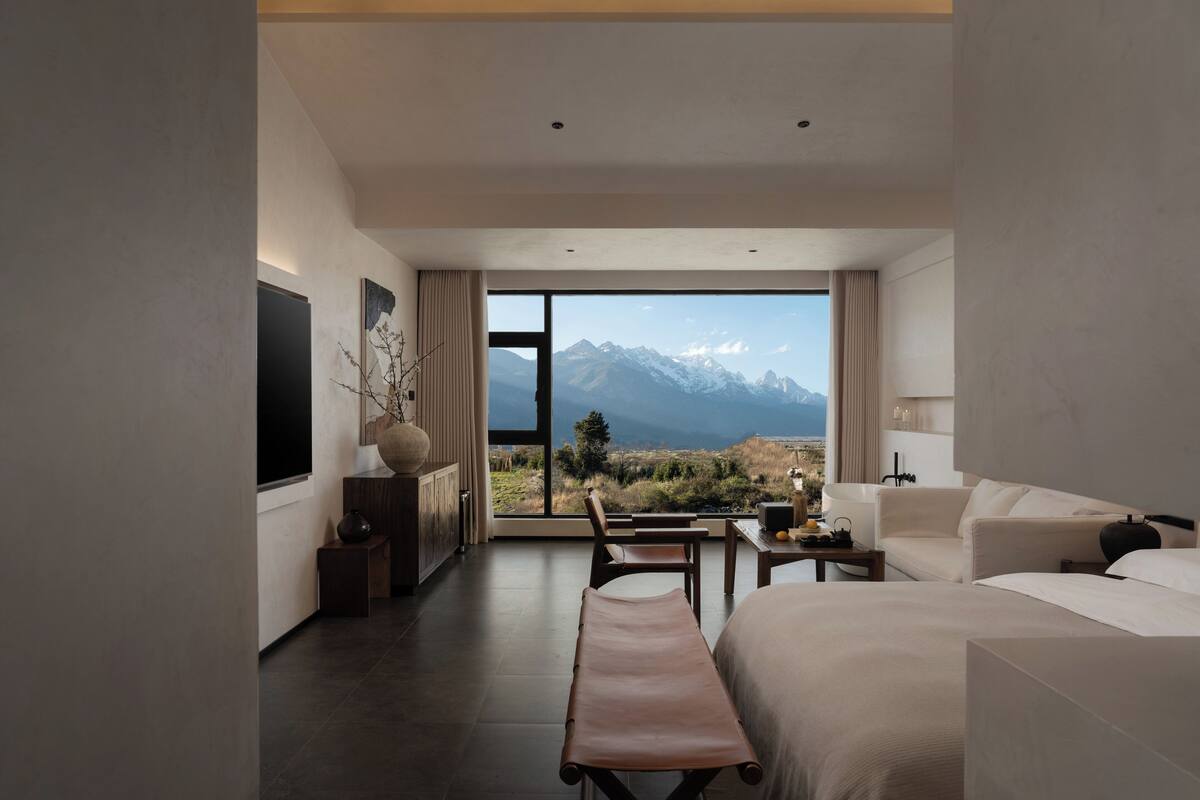 A modern hotel room with a large bed, a bench, a desk, and a view of mountains.