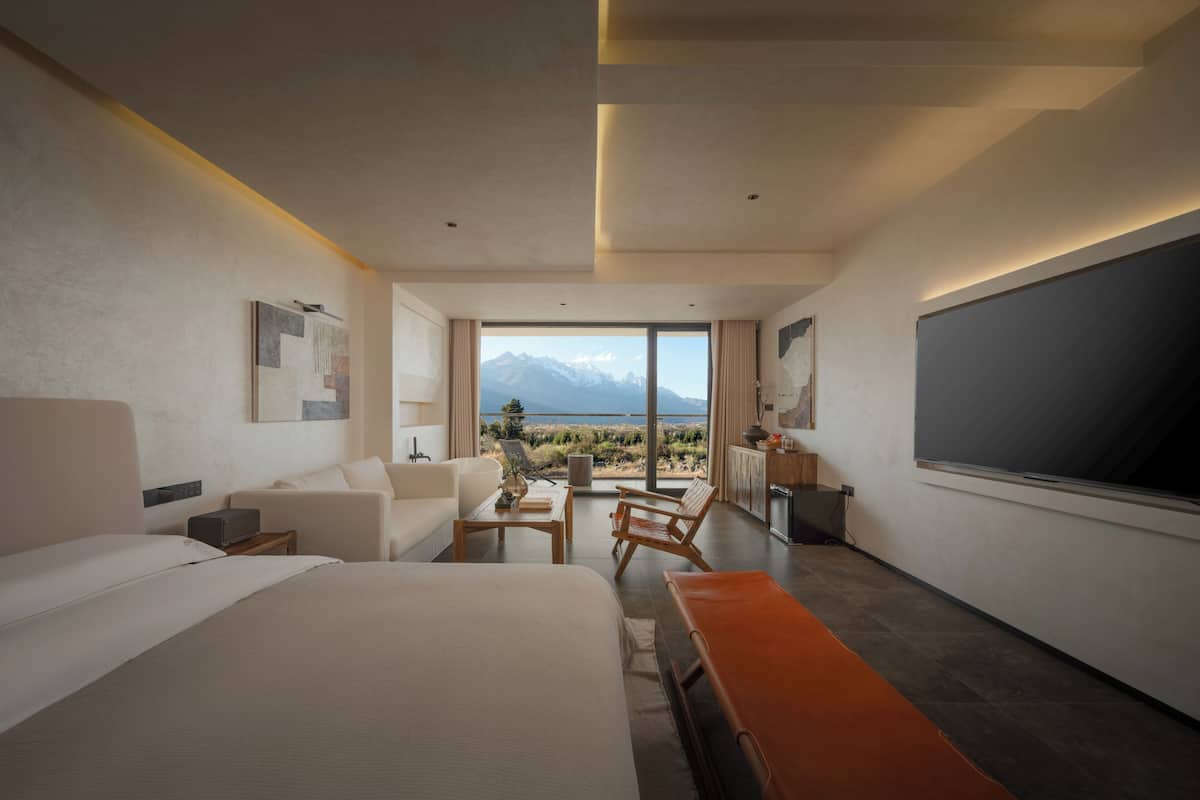 A modern hotel room with a large bed, a desk, a sofa, and a view of mountains.