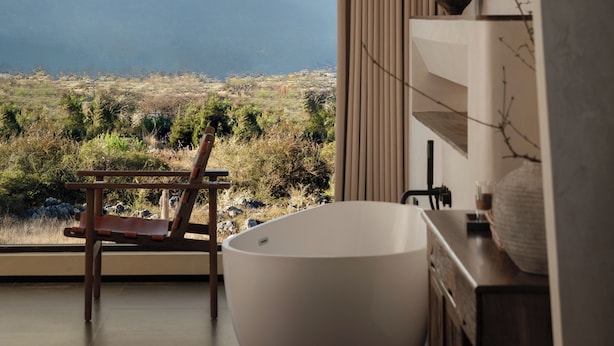 A modern bathroom with a freestanding bathtub, a wooden chair, and a view of mountains through a large window.