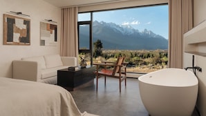 A modern hotel room with a large window offering a mountain view, a bathtub, a sofa, and a coffee table.