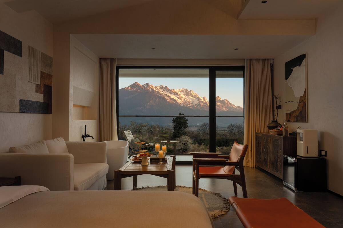 A hotel room with a large window offering a mountain view, a bed, a sofa, a dining table with candles, and a cabinet.