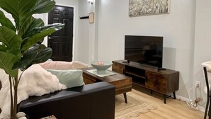 A living room with a black sofa, a coffee table, a TV on a wooden stand, a large plant, and a painting on the wall. - Stylish Studio in Old Colorado City-Shops and Dining Only Steps Away! (Colorado Springs)