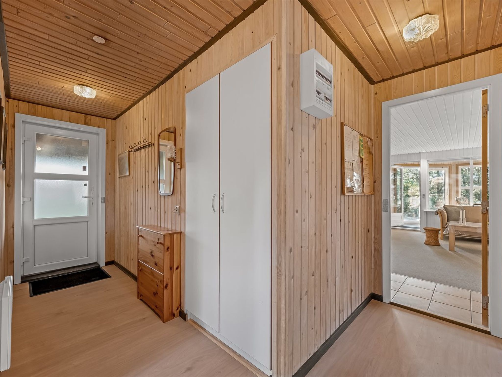 A wooden-paneled room with a white door, a mirror, a wooden cabinet, and a bulletin board.