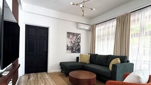 Un salon moderne avec un canapé d’angle, une table basse ronde, un téléviseur à écran plat et un luminaire suspendu au plafond.
