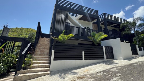 Une maison moderne à deux étages, avec un escalier menant à l’entrée, entourée de verdure et sous un ciel dégagé.