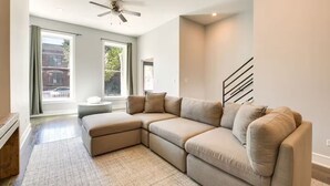 A modern living room with a sectional sofa, a ceiling fan, and large windows offering a view of the street. - NEW! Stylish 3BD/2.5BTH w/parking! United Center! (Chicago)