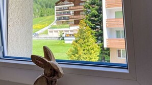 Ikkunasta avautuu näkymä rakennukseen, jossa on parvekkeita, ja taustalla kohoaa vuori.