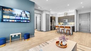 A modern living room with a flat-screen TV displaying a movie, a kitchen with a dining area, and a hallway leading to bedrooms.