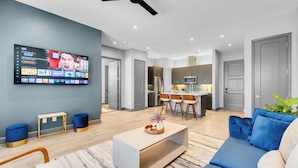 A modern living room with a TV, sofa, coffee table, and a dining area with chairs and a kitchen in the background.