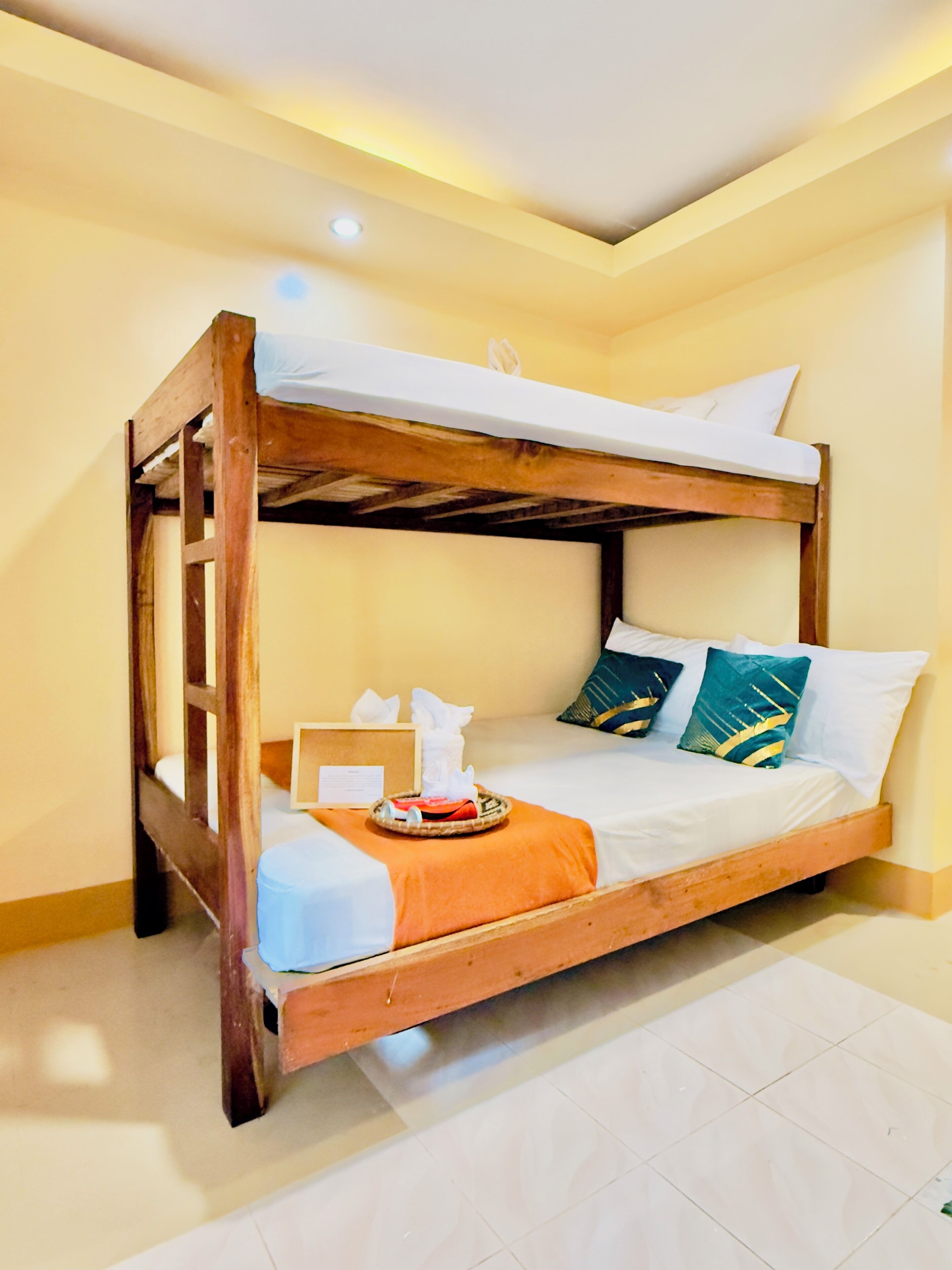 Una cama litera con estructura de madera, ropa de cama blanca y almohadas decorativas. Hay una bandeja con un pañuelero y un pequeño objeto sobre la cama.