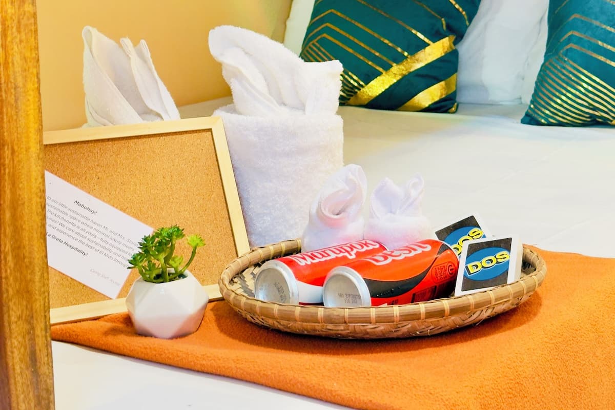 Una cama bien tendida con almohadas blancas y turquesa, una cesta con latas y una tarjeta, y una pequeña planta en maceta sobre un cabecero de madera.