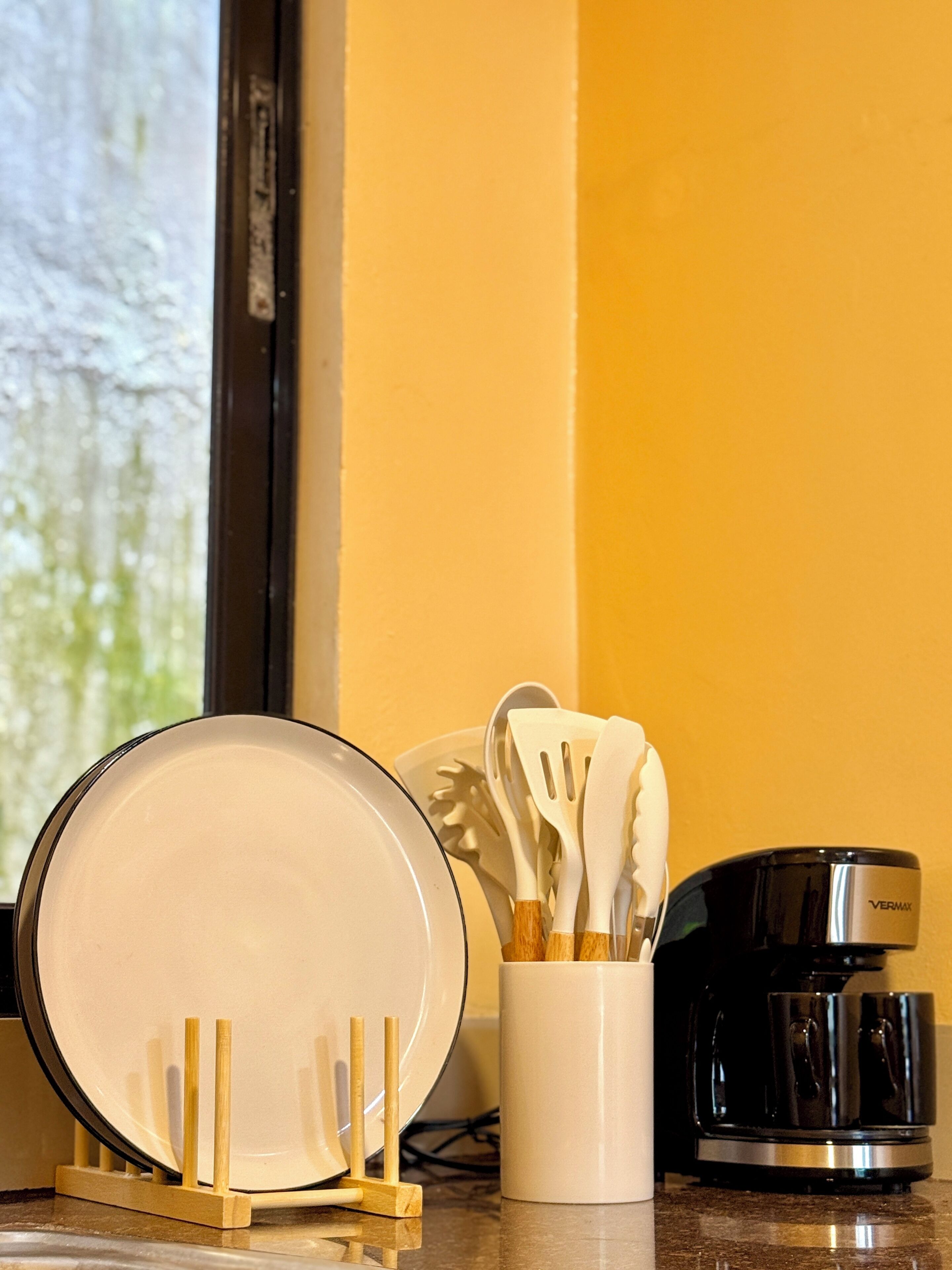 Una encimera de cocina con un plato blanco, una taza blanca, una cafetera negra y un juego de utensilios blancos.