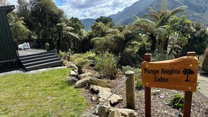 A wooden sign for Punga Heights Cabin with a palm tree symbol, surrounded by a garden with rocks and ferns, and a staircase leading to a building.