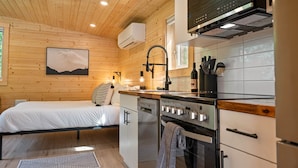 A compact bedroom with a bed and bedside table, a small kitchenette with a stove and sink, and a wooden ceiling. - Arctic Lodge - Alpha Mountain Loding (Revelstoke)
