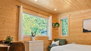 A wooden-paneled room with a bed, a desk with a chair, a dresser, and two windows offering a view of trees. - Arctic Lodge - Alpha Mountain Loding (Revelstoke)