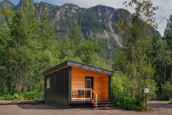 A small cabin with a wooden exterior and a porch, surrounded by dense forest and mountains. - Arctic Lodge - Alpha Mountain Loding (Revelstoke)