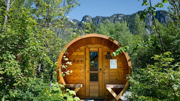 A wooden sauna structure with a door and benches, surrounded by lush greenery and mountains in the background. - Arctic Lodge - Alpha Mountain Loding (Revelstoke)