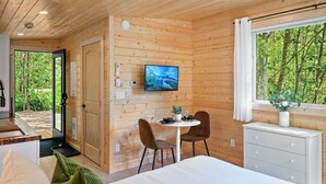 Ein Holzblockhaus-Interieur mit einem Bett, einem Esstisch mit Stühlen, einem an der Wand befestigten Fernseher und einem Fenster mit Blick auf Bäume.