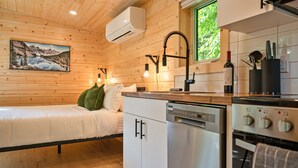 Ein Interieur einer Holzhütte mit einem Bett, einer Küche mit modernen Geräten und einem Fenster mit Blick auf Grünflächen.