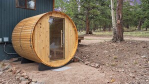 A wooden sauna structure with a glass door, situated in a forested area with a house in the background.