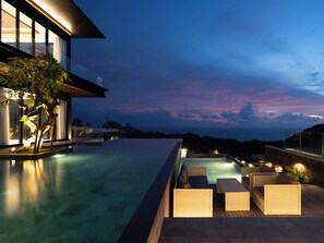 Una piscina infinita moderna con asientos exteriores, una estructura de vidrio y vista al océano al anochecer.
