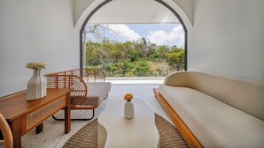 Una sala de estar moderna con un sofá, una mesa de madera y una silla. Hay un ventanal que ofrece vistas a árboles y colinas.