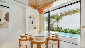 Un comedor moderno con una mesa de vidrio y sillas de madera, un accesorio de iluminación decorativo y vistas a una piscina.