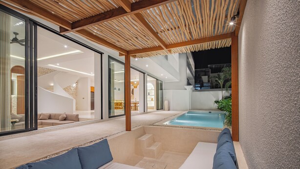 Un área de piscina moderna al aire libre con una pérgola de madera, una zona de estar con cojines y una escalera que lleva a un nivel superior.