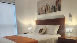 A modern hotel room with a large bed, two bedside tables, a nightstand, and a cityscape painting on the wall. - A HOME AWAY FROM HOME (Lima)