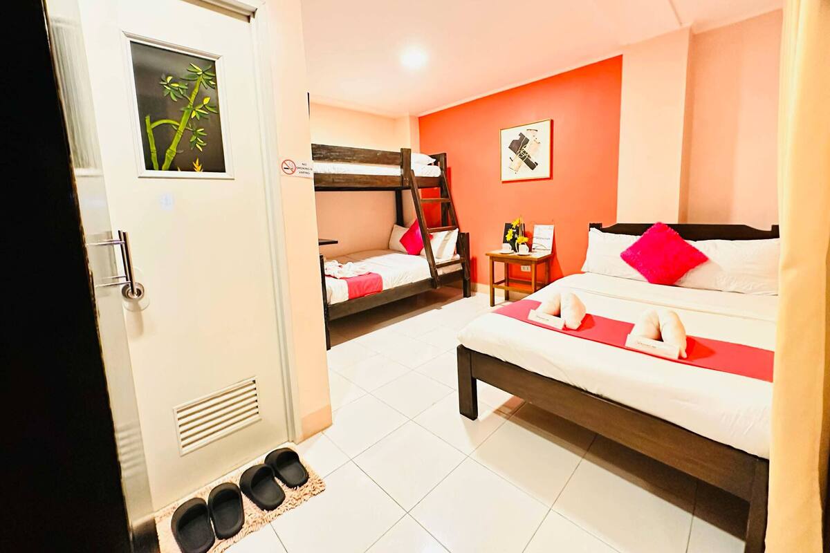 Une chambre d’hôtel avec un lit superposé, un lit simple, une petite table avec un vase et des chaussons sur le sol.