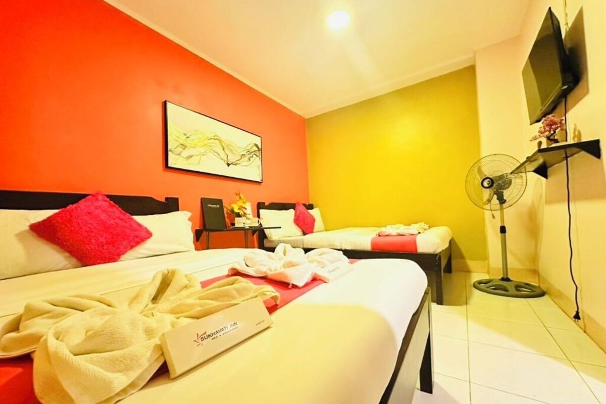Une chambre d’hôtel avec deux lits, un tableau au mur, un téléviseur fixé au mur et un ventilateur sur pied.