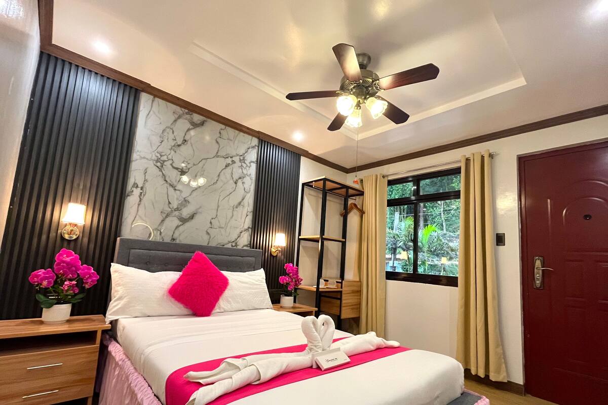 Une chambre d’hôtel avec un lit, un ventilateur de plafond, une fenêtre avec des rideaux, une porte rouge et un mur orné d’un motif rappelant le marbre.