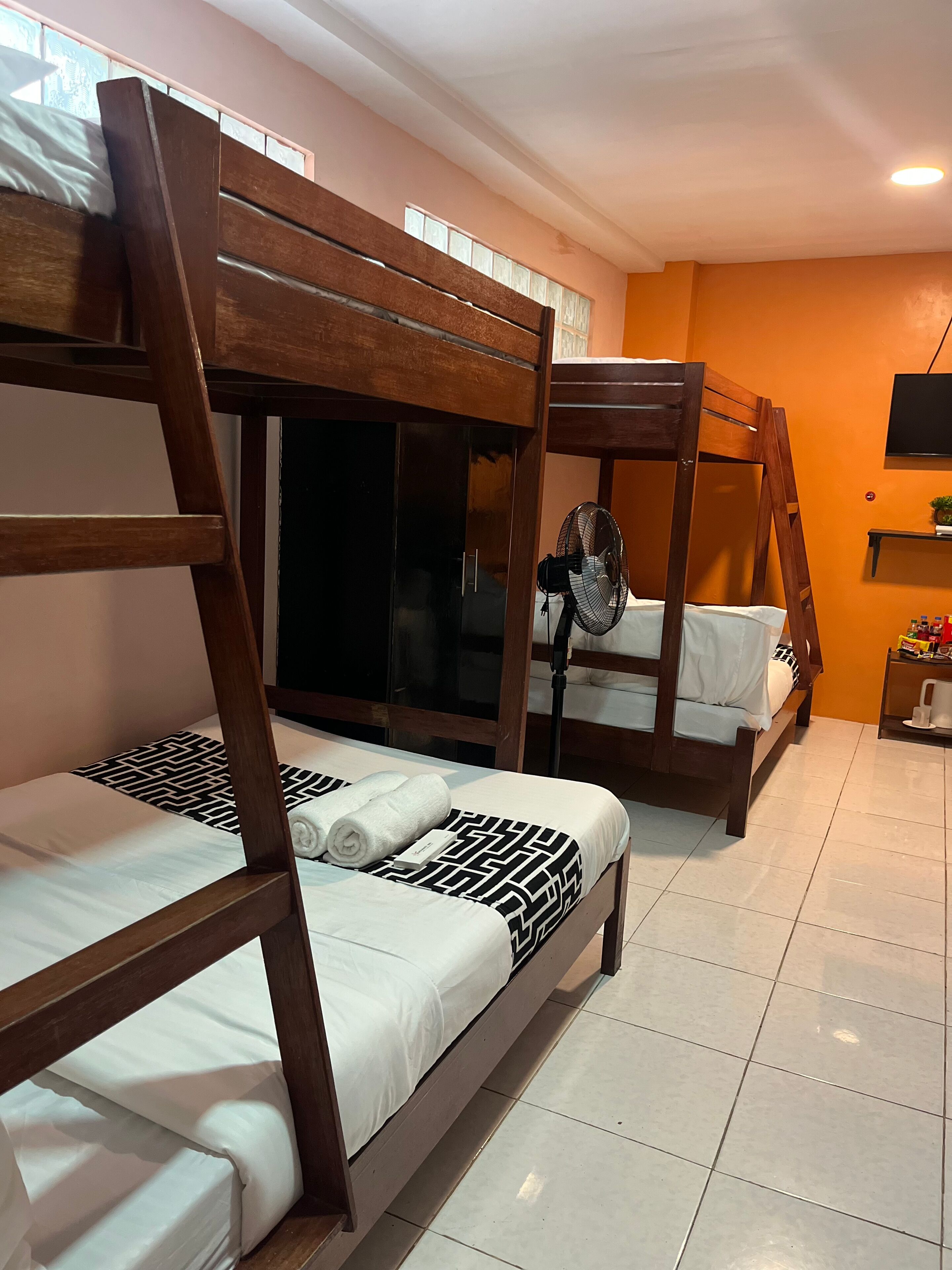 A room with two bunk beds, a fan, a TV on the wall, and a small shelf with items.