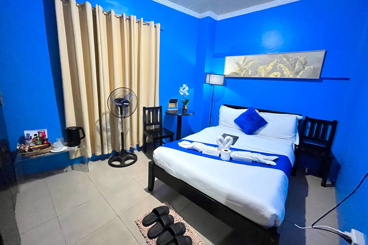 Une chambre d’hôtel comprenant un lit, un ventilateur, une chaise, une petite table avec une lampe et une œuvre d’art accrochée au mur.