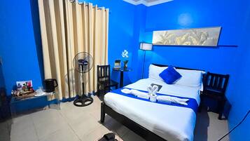 A hotel room with a bed, a fan, a chair, a small table with a lamp, and a wall-mounted artwork.